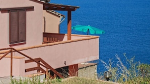 Une maison située sur la côte, dotée d’un balcon donnant sur la mer, d’un escalier en bois et d’un parasol vert.