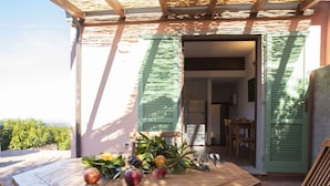 Une table en bois, ornée de fruits et d’un vase de fleurs, se trouve sur une terrasse aux volets verts, d’où l’on peut admirer la mer.