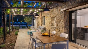 Un patio al aire libre con pared de piedra, pérgola de madera y luces colgantes. Hay mesas y sillas, y un pequeño jardín con plantas y un árbol.