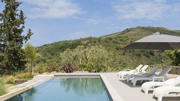 Una piscina con sillas de descanso y una sombrilla grande, rodeada de vegetación y colinas.