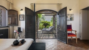 Una habitación con un umbral abierto que da a una zona de estar al aire libre, una cama con una bandeja de té y tazas, y una silla con un cojín colorido.