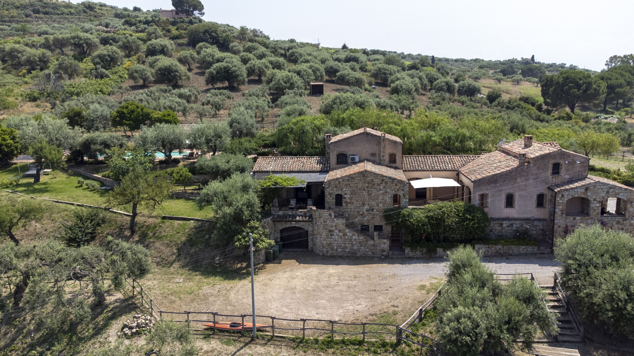 A stone building with a courtyard, surrounded by olive trees and a hilly landscape.