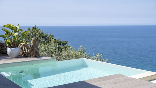A rectangular infinity pool with clear blue water, surrounded by a stone wall and potted plants, overlooking a calm sea.