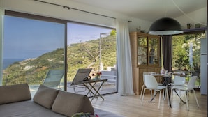 A modern living room with a large sliding glass door leading to a balcony with a scenic view of a hillside and the sea.