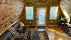 A cozy attic room with wooden walls, a black leather sofa, a small table, and a large window. - The Lookout | Secluded A-Frame | Hot Tub | Games (Campton)