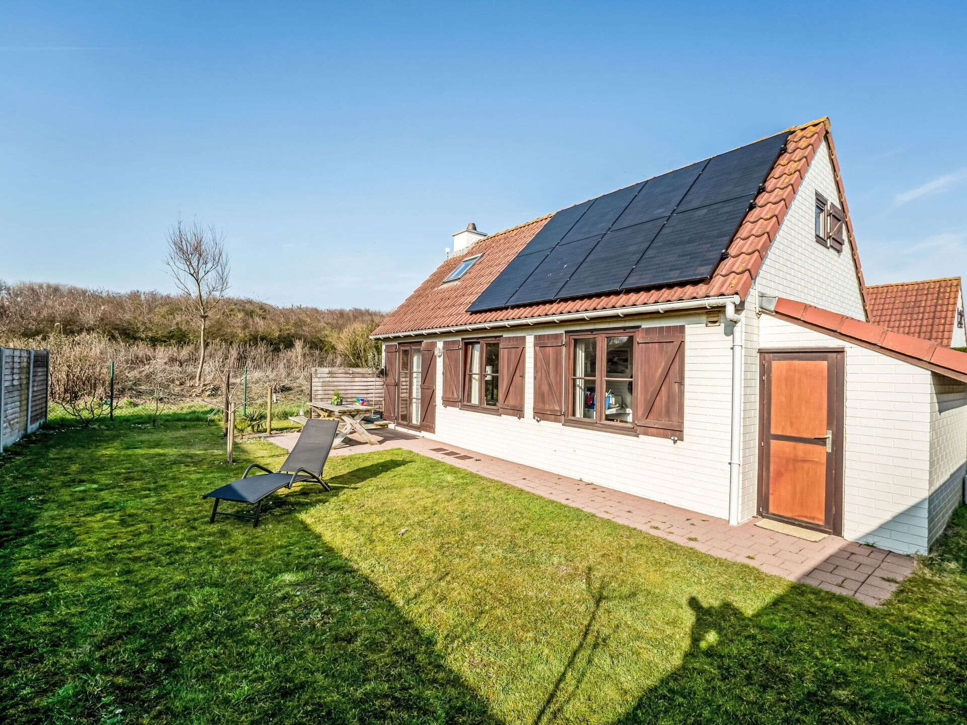 Ein Haus mit geneigtem Dach, Solaranlagen, einer Holztür und einem Rasen mit einer Liege.