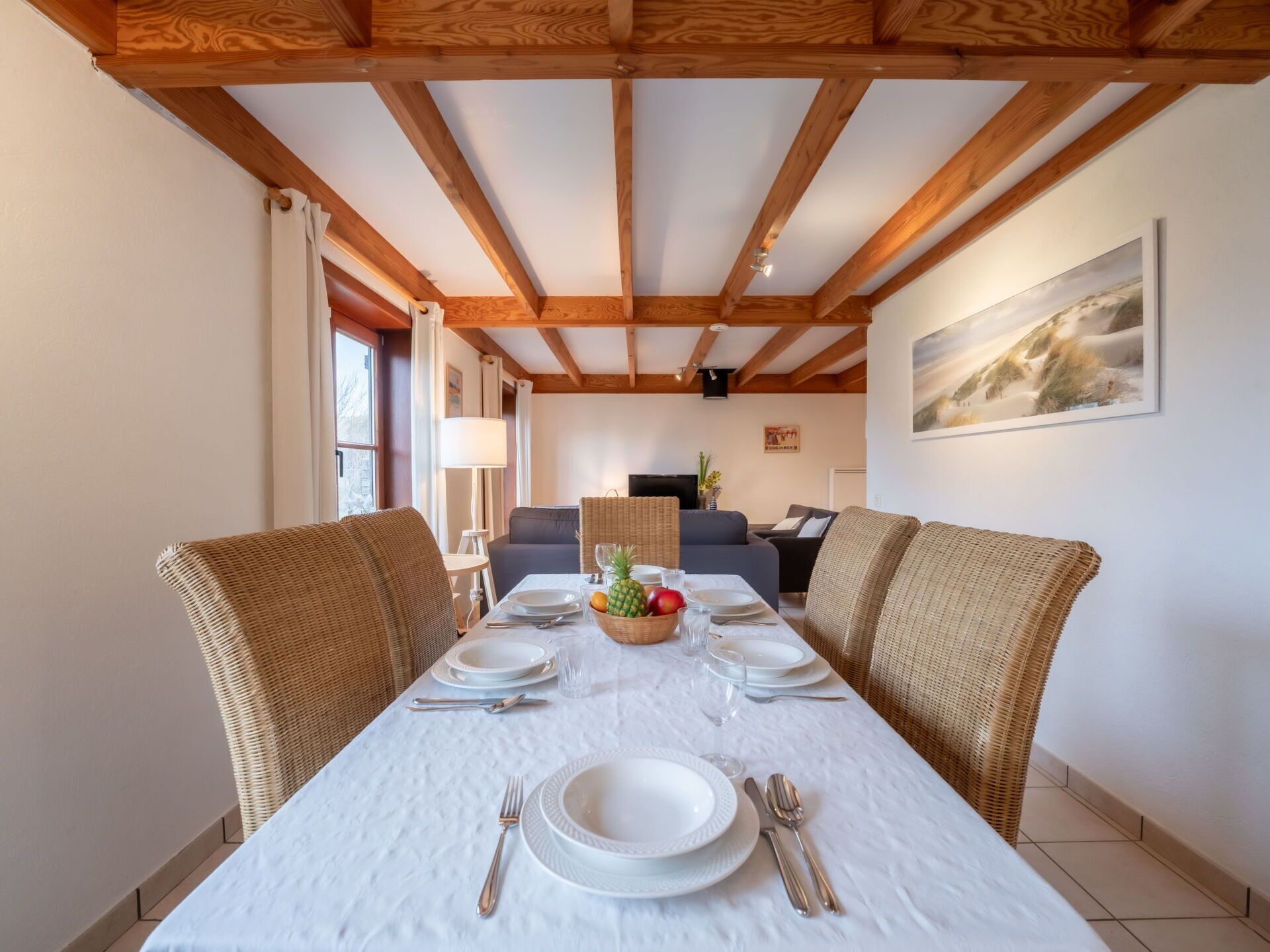 Uma sala de jantar com uma mesa comprida posta para quatro pessoas, rodeada por cadeiras de vime, teto de madeira e um quadro na parede.