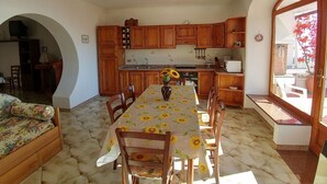 Un comedor con una mesa dispuesta para seis personas, rodeada de sillas de madera. Se ve una cocina con armarios de madera y una encimera blanca al fondo.