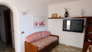 Una sala de estar con un sofá, un televisor de pantalla plana, un estante montado en la pared con objetos decorativos y suelo de baldosas.