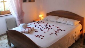 Una cama bien tendida con pétalos de rosa, una toalla enrollada y una lámpara en una mesita de noche en una habitación con ventana y un cuadro en la pared.