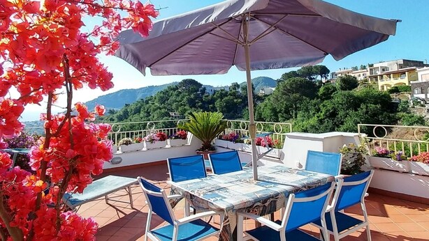 Un patio al aire libre con una mesa y sillas, un paraguas grande y vistas a una colina con edificios.