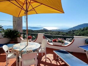 Eine Terrasse mit Tisch und Stühlen, einem gelben Sonnenschirm und Blick auf das Meer und die Berge.