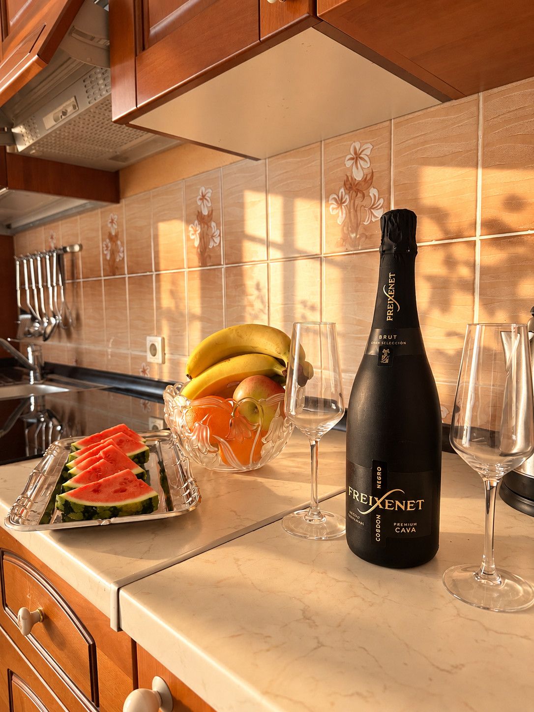 Een keukentafel met een fles Brut Cava, een schaal fruit en twee wijnglazen.