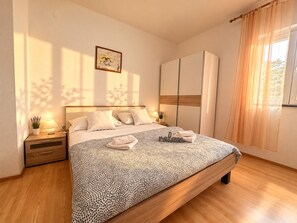 A modern bedroom with a large bed, wooden nightstands, a bedside table with a lamp, and a window with sheer curtains. - Apartment Bodulo 2 (Povljana)