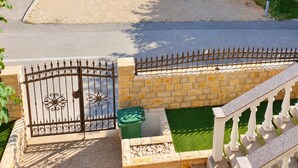 Zona residencial con entrada porticada, camino de entrada pavimentado y un pequeño jardín con contenedor verde y banco blanco.