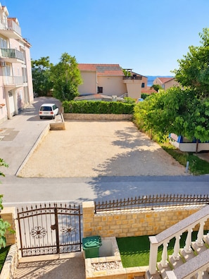 Bairro residencial com calçada pavimentada, entrada com portão e pequena área de jardim com um recipiente verde e um banco branco.