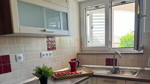 Une cuisine moderne avec des meubles blancs, un four à micro-ondes, une fenêtre avec des stores et un plan de travail agrémenté d’une plante et d’un plateau de fruits coupés.