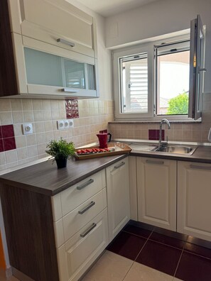 Una cocina moderna con armarios blancos, un microondas, un ventanal con persianas y una encimera con una planta y una bandeja de fruta cortada.