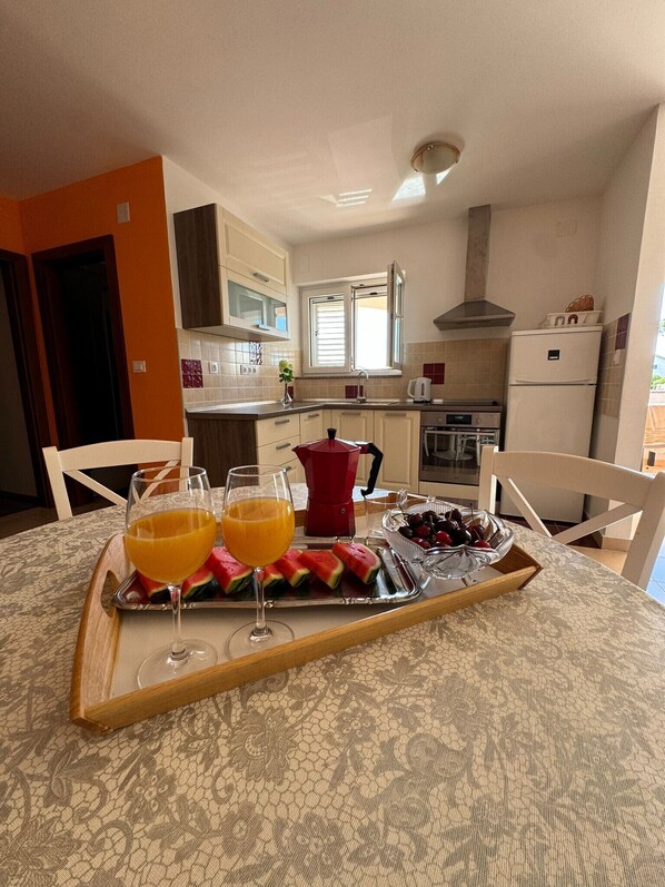 Un comedor con una mesa preparada para dos personas, que incluye una bandeja con un vaso de jugo de naranja, un vaso de agua, rebanadas de sandía y un tazón de cerezas.