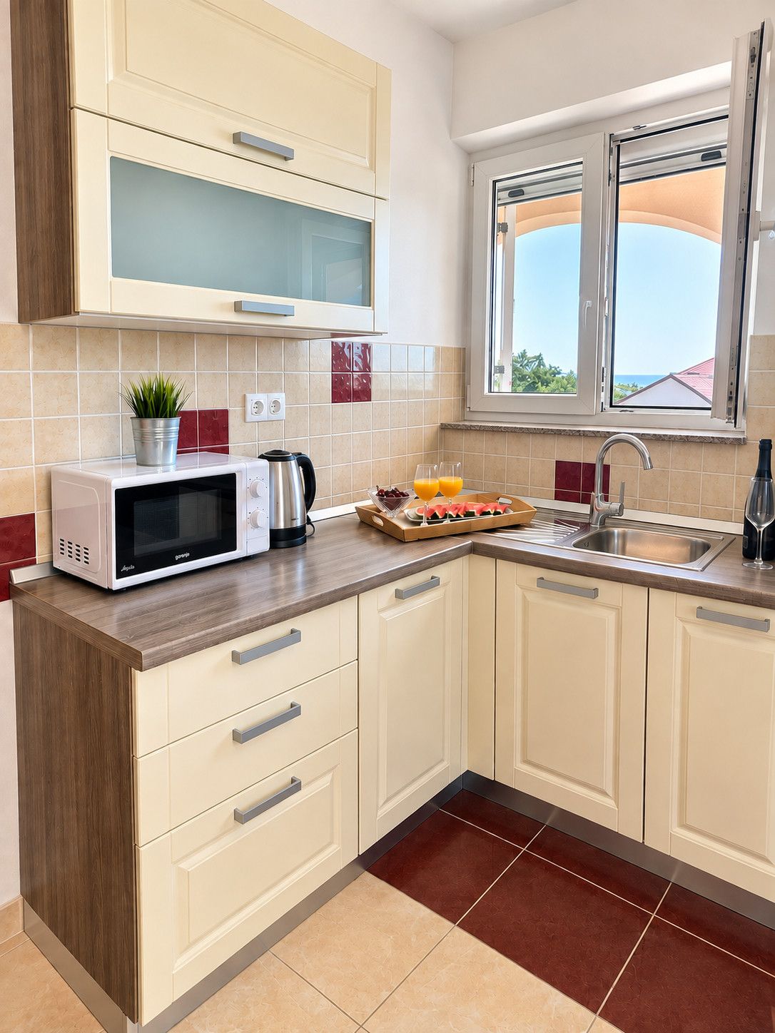 A kitchen with a microwave, a kettle, a fruit tray, and a window with a view.