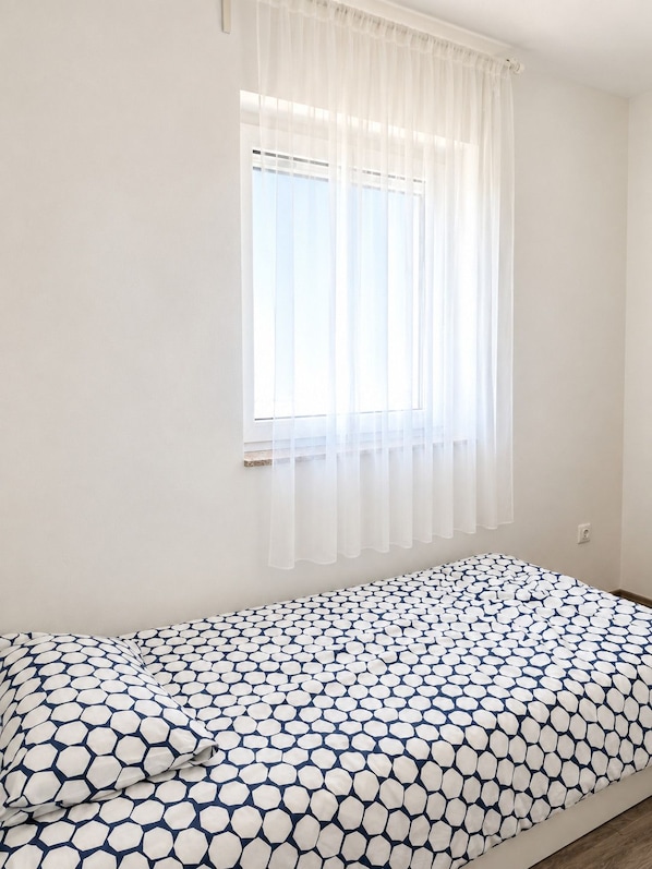 A room with a single bed, a window with sheer curtains, and a plain white wall.