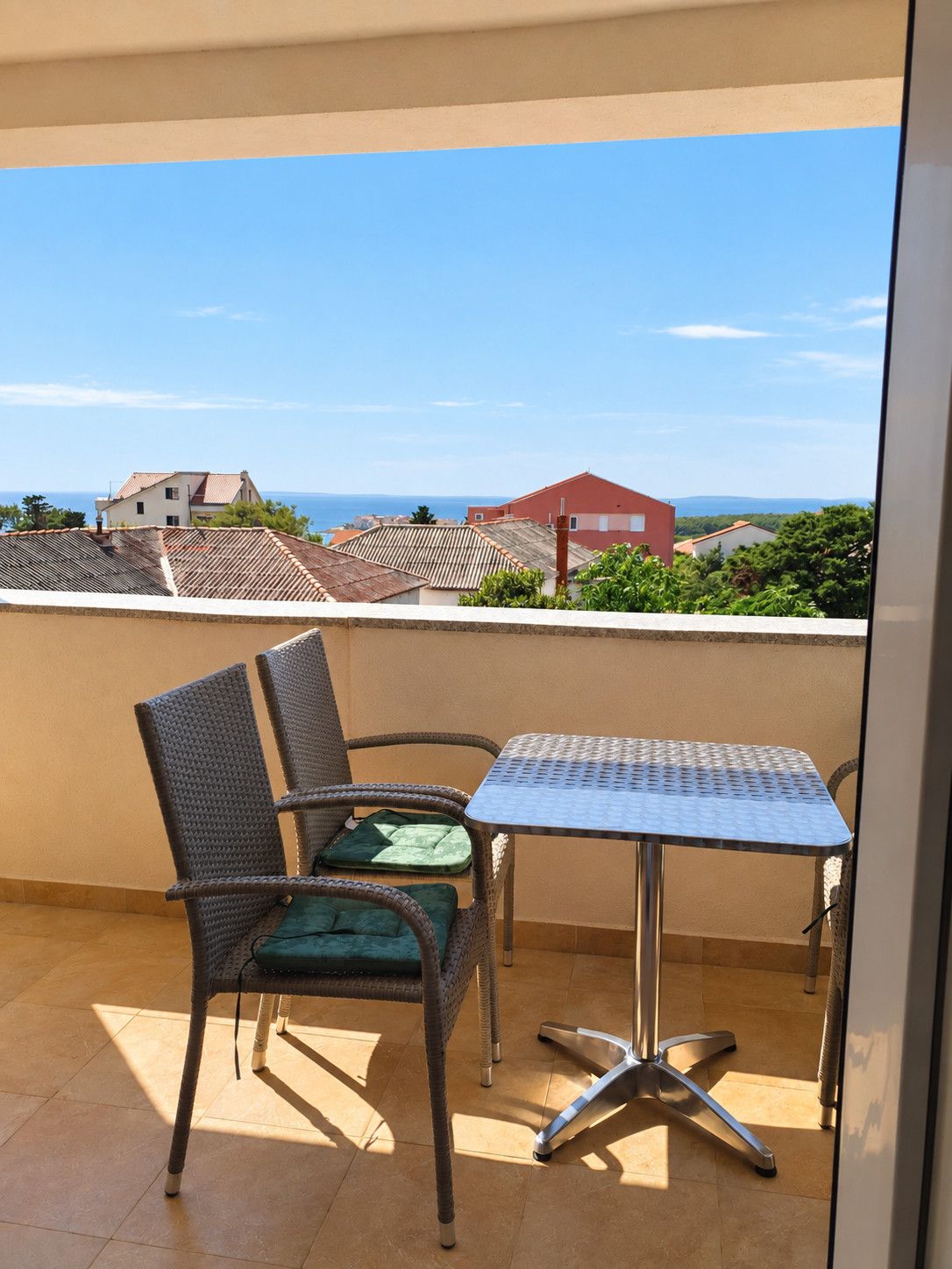 Varanda com mesa de metal e cadeiras de vime, com vista para uma área residencial com casas e céu claro.