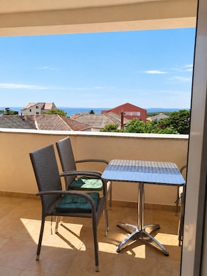 A balcony with a metal table and wicker chairs, overlooking a residential area with houses and a clear sky.