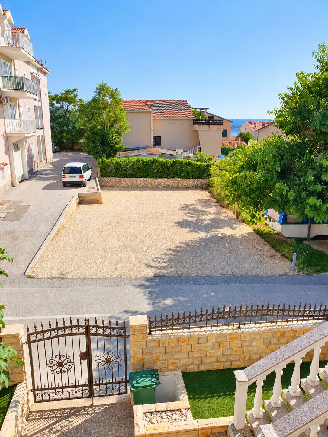 Zona residencial con entrada porticada, camino de entrada pavimentado y un pequeño jardín con contenedor verde y banco blanco.
