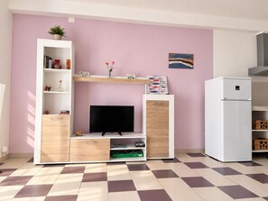 A living room with a flat-screen TV, wooden entertainment unit, pink wall, and a white refrigerator.