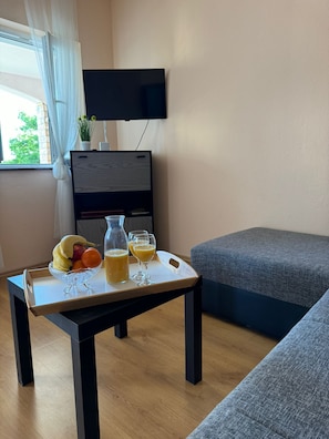 Uma pequena sala de estar com uma mesa de madeira com frutas e suco, um sofá cinza e uma TV na parede.