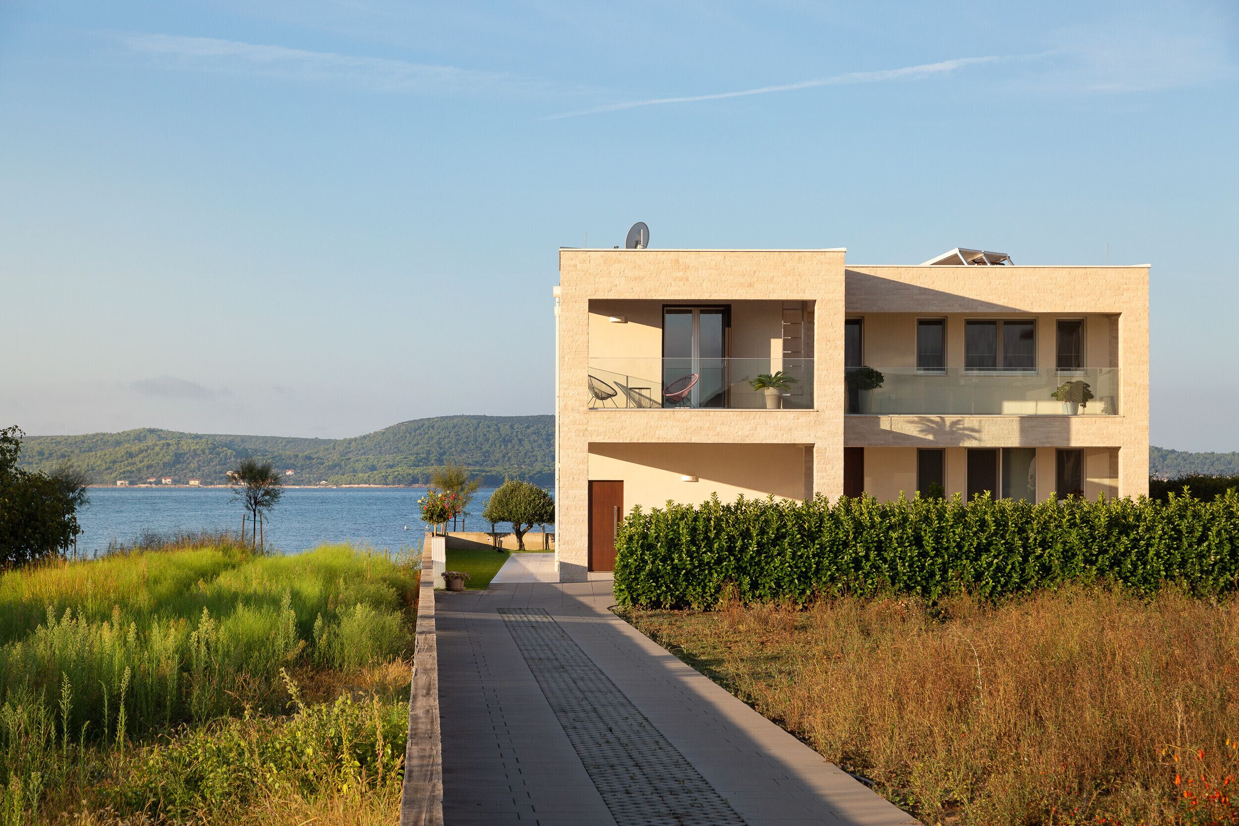 A modern two-story building with a balcony, a paved walkway, and a view of a body of water and hills in the background.