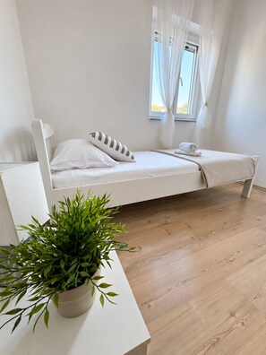 Ein Einzelbett mit weißem Kopfteil, ein Nachttisch mit Topfpflanze und ein Fenster mit transparenten Vorhängen.