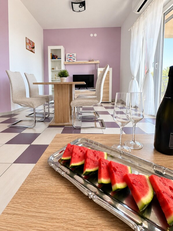 A modern living room with a dining area, featuring a checkered floor, a dining table with chairs, a TV, and a glass tray with watermelon slices.