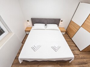 A neatly made bed with white linens and two folded towels, a grey upholstered headboard, bedside tables with lamps, and a wooden floor.