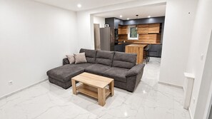 Un salon moderne avec un canapé d’angle, une table basse en bois et une cuisine en arrière-plan.