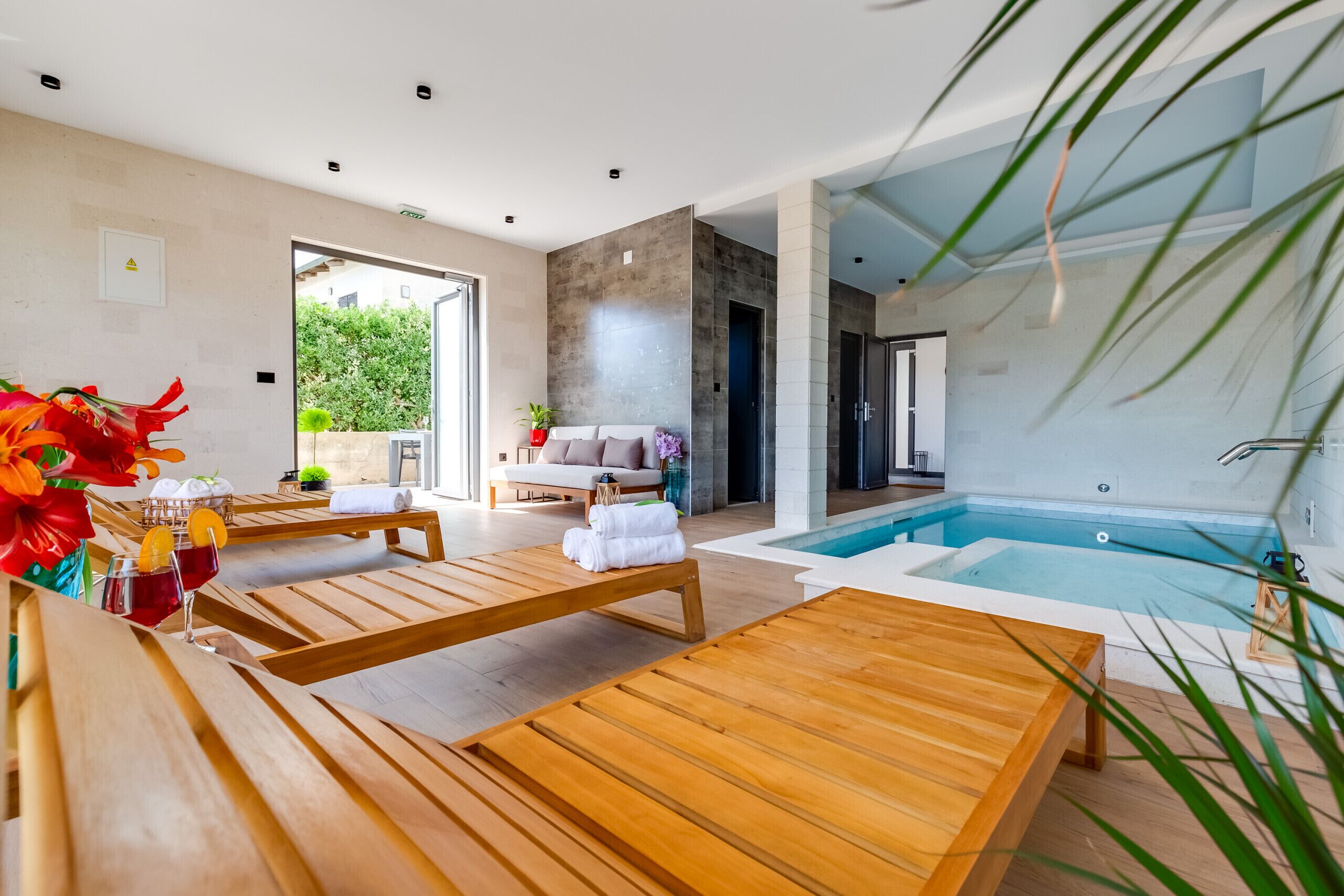 Un espace spa moderne doté de chaises longues en bois, d’une piscine et d’un coin salon offrant une vue sur la verdure environnante.