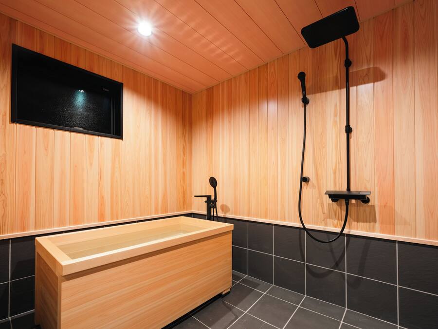 A modern bathroom with wooden panel walls, a wooden bathtub, black tiled flooring, and a mounted television.