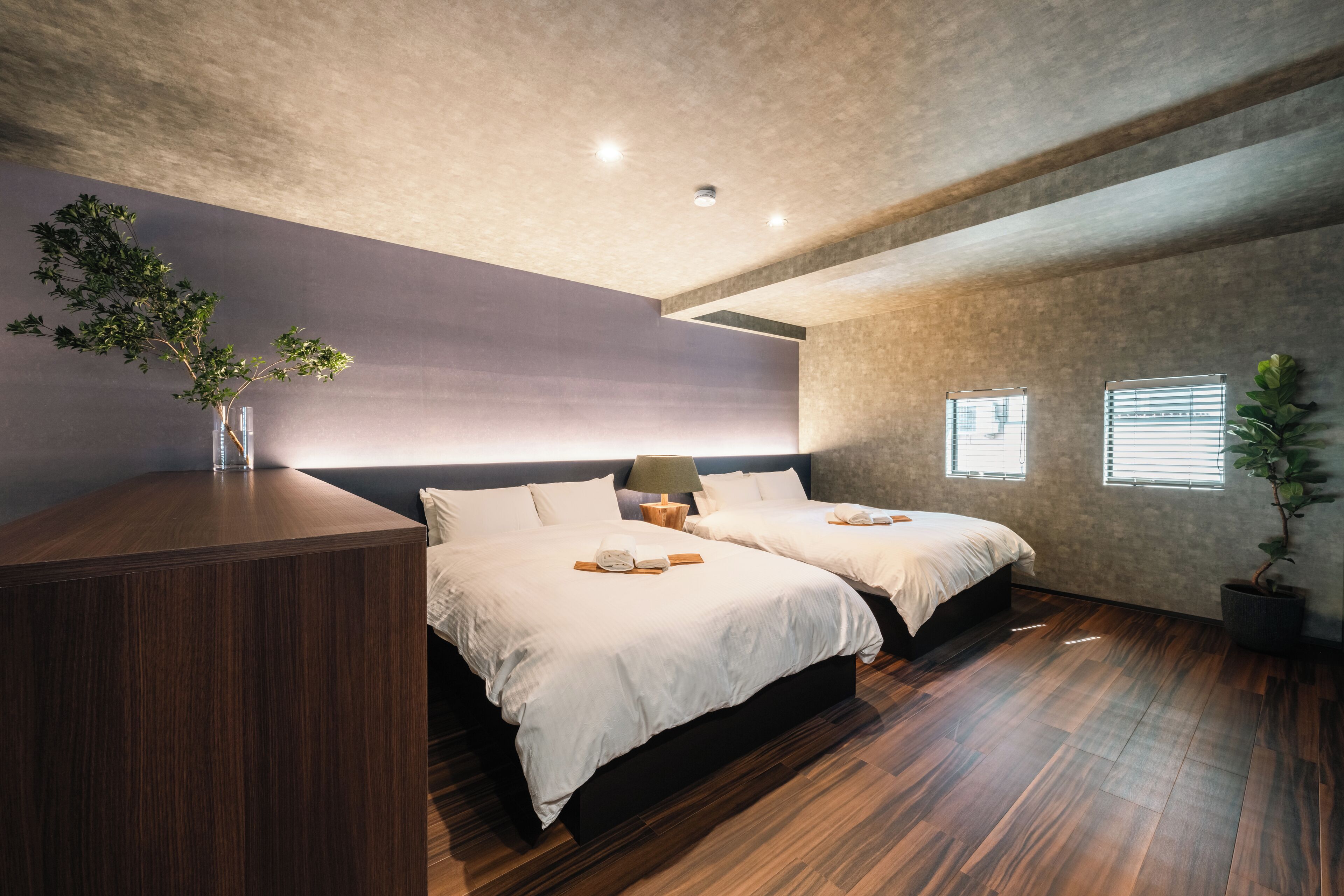 A modern bedroom with two beds, a wooden dresser, a potted plant, and a large window with blinds.
