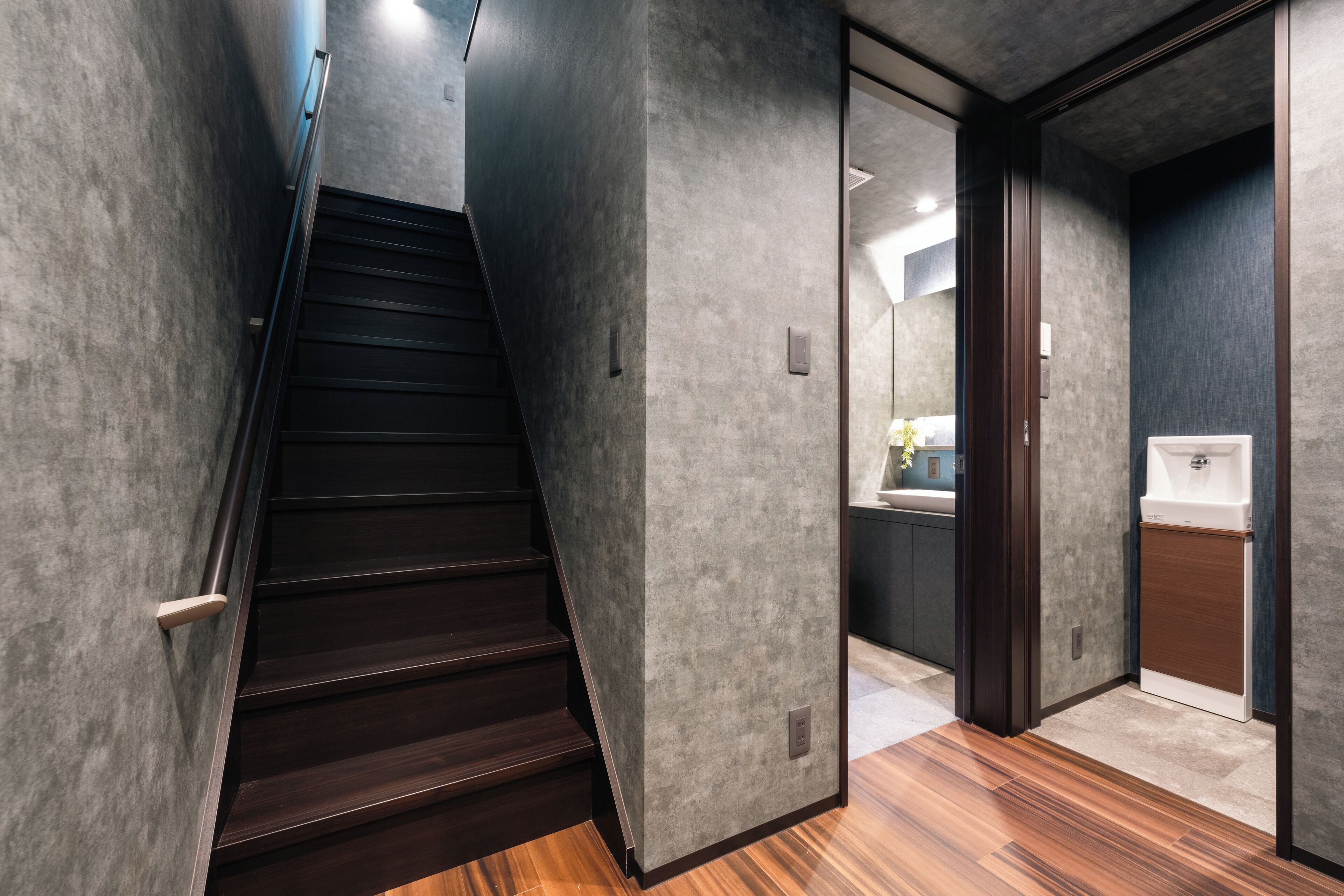 A modern interior with a wooden staircase, grey textured walls, and a bathroom with a white sink and mirror.