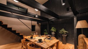 Un comedor con una mesa de madera preparada para cuatro, rodeada de sillas de madera. En el fondo, se ve una escalera con vigas de madera oscura.