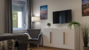 A modern living room with a grey sofa, a white cabinet with gold handles, a flat-screen TV mounted on the wall, a floor lamp, and a potted plant.