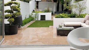 A modern outdoor patio area with a glass wall, a white sofa, a glass coffee table, and a small garden with a lawn and potted plants. - Softlife Apartments  (Abuja)