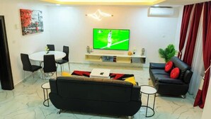 A living room with a flat-screen TV, a black sofa, a dining table with chairs, and a potted plant. - Softlife Apartments  (Abuja)