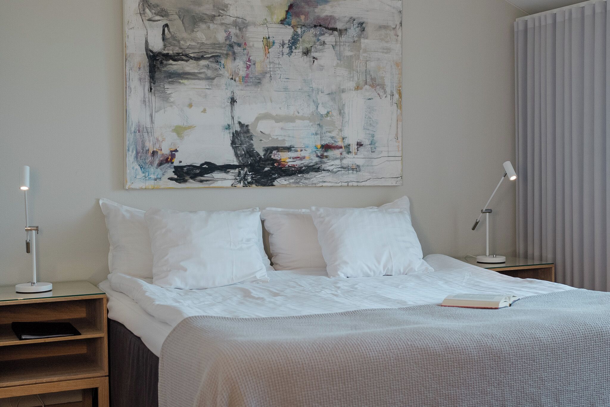 Un dormitorio moderno con un cuadro abstracto grande, una cama bien hecha con ropa de cama blanca y dos lámparas de noche.