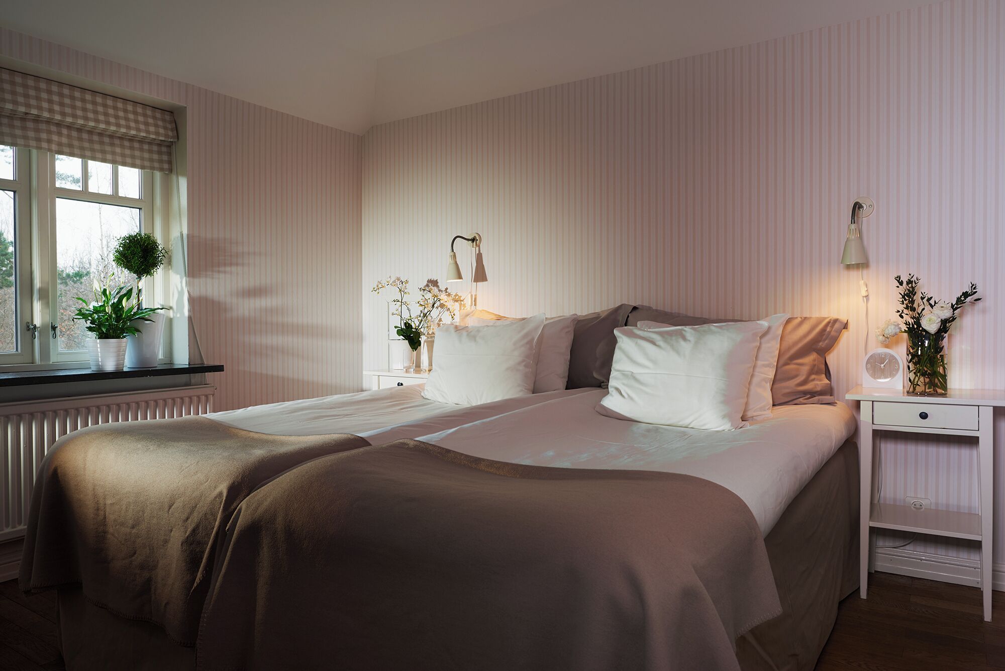 A bedroom with two beds, a window with curtains, a bedside table with a lamp, and a small plant on the windowsill.