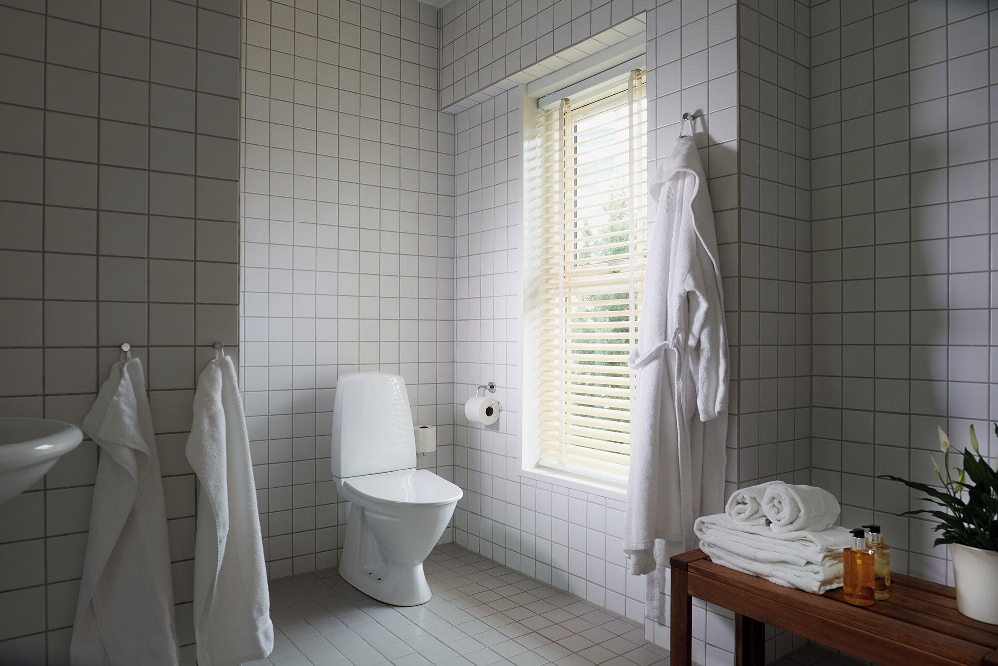 Un baño con paredes de azulejos blancos, un inodoro, un ventanal con persianas y una mesa de madera con toallas y artículos de aseo.