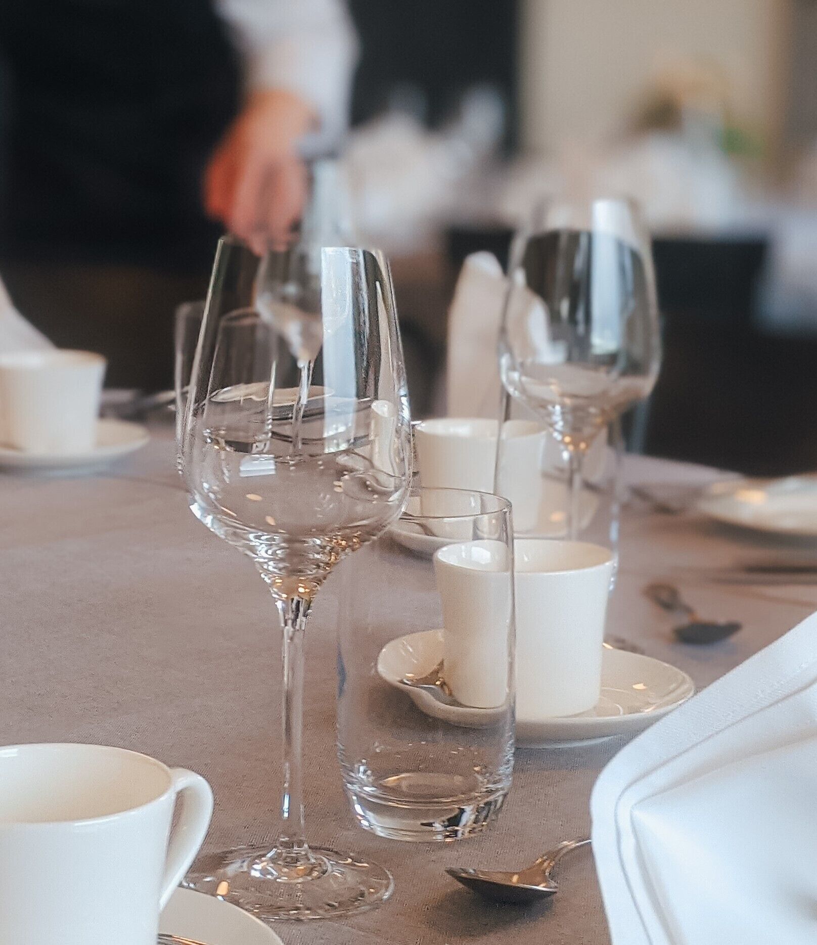 Una mesa dispuesta con copas de vino transparentes, tazas y platillos blancos, evocando un ambiente de comedor formal.