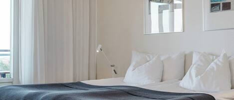 A neatly made bed with white linens and a dark blue blanket, a bedside lamp, and a wall with a framed map and sketches.