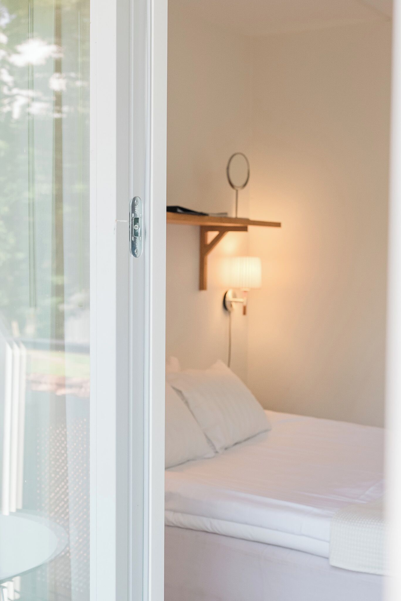 A bedroom with a bed, a bedside table, a lamp, and a window with a view of greenery.