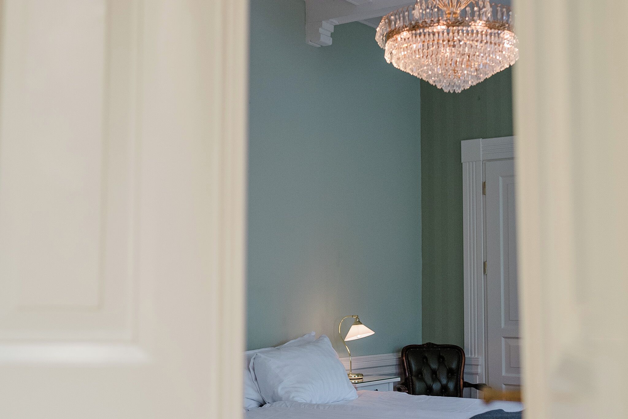 A hotel room with a bed, a nightstand, a lamp, a chair, and a chandelier.